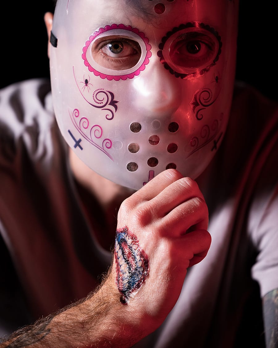 En person som bär en dekorerad vit mask med rosa och svarta mönster håller en knytnäve mot ansiktet och avslöjar ett falskt blodigt sår skapat med tatuajes falsos para Halloween. Den dramatiska belysningen framhäver både masken och den realistiska skadan.
