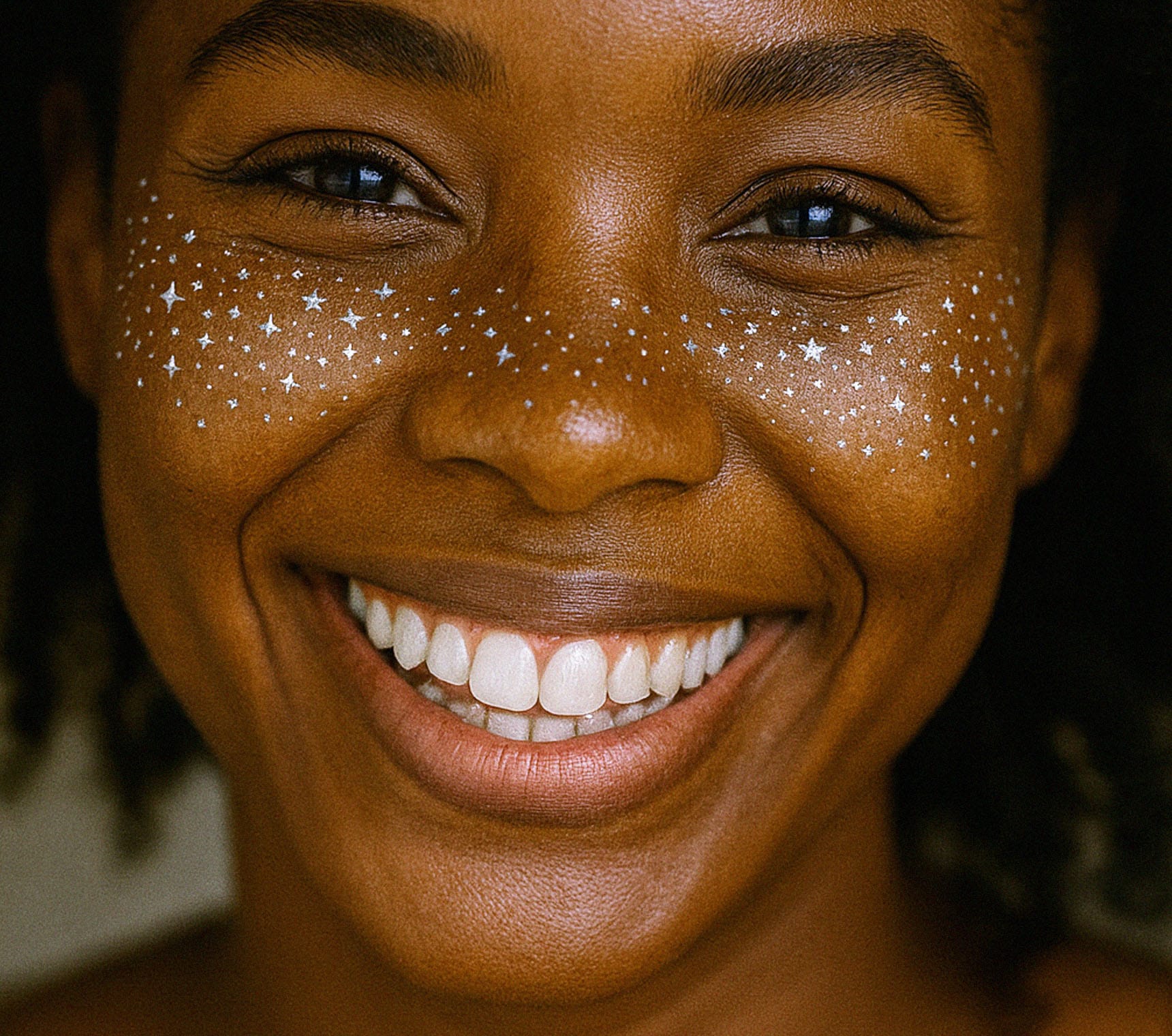 A smiling person with brown skin and curly hair wears Freckle Tattoos Silver Stars across their cheeks and nose, creating a playful, whimsical look with tiny silver star designs that resemble enchanting freckle art.
