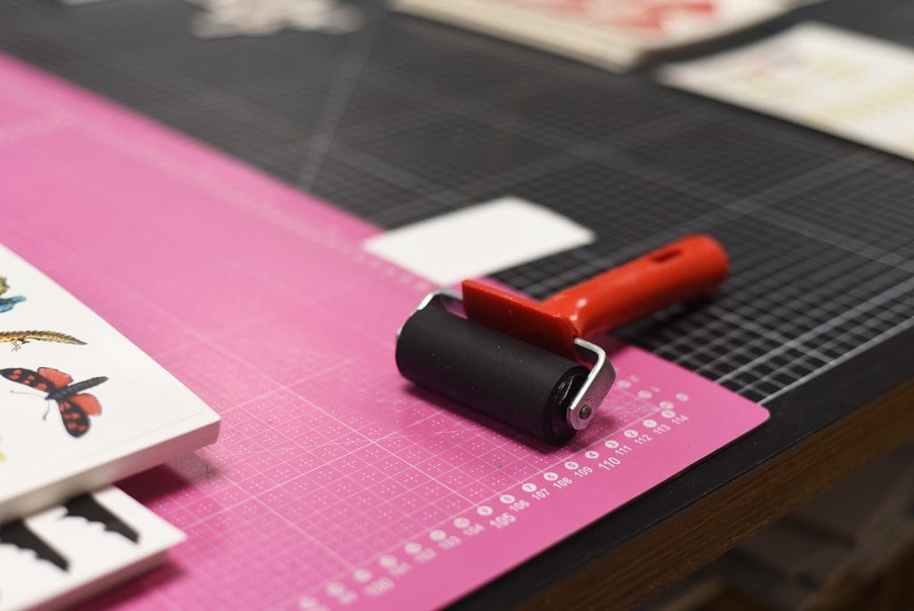 En brayer roller med rött handtag vilar på en rosa skärmatta med måttmarkeringar. Färgade illustrationer, tilpassede midlertidige tatoveringer och pysselmaterial syns i bakgrunden på ett svart rutmönstrat bord.