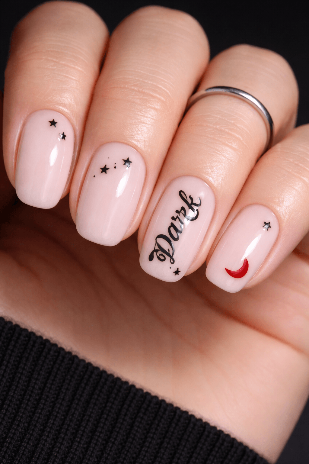 A hand with pale pink manicured nails featuring small black stars, the word Dark in black script, and a red crescent moon design. The person wears a silver ring on their middle finger.