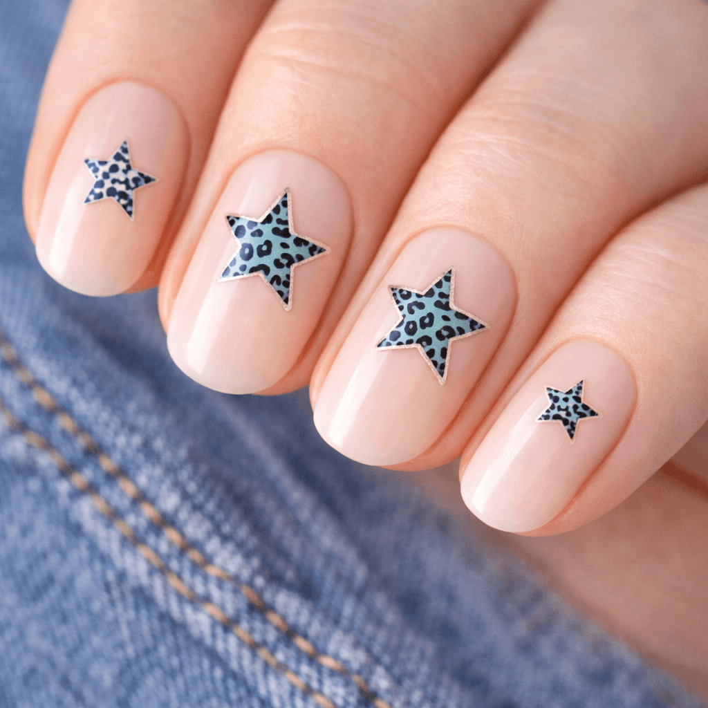 A close-up of a hand with nude-colored nail polish, each nail decorated with a star-shaped design featuring a blue and black leopard print pattern. The hand rests on denim fabric.