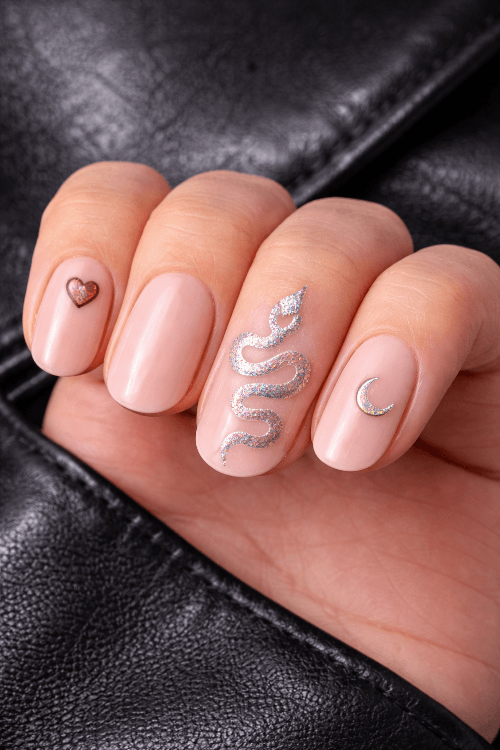 A hand with nude-pink manicured nails featuring small designs: a glittery pink heart, a shimmering silver snake, and a silver crescent moon. The background is black leather.