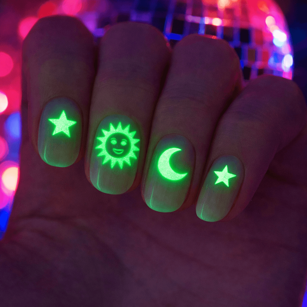 A close-up of four fingernails glowing under blacklight, each decorated with a neon green star, sun with a face, crescent moon, and another star design. The background is blurred with colorful lights.