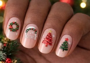 A close-up of four fingernails with festive Christmas nail art, including a wreath, the word Xmas, and two decorated Christmas trees on a nude base, with small white snowflakes on each nail.