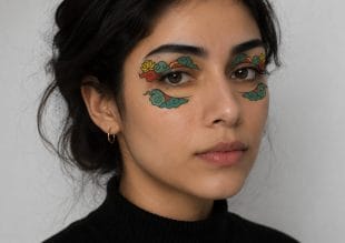 A woman with dark hair and a black polo neck poses against a plain background. She has colourful, artistic designs resembling clouds and flowers painted around her eyes.
