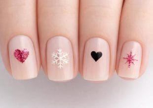 Close-up of four fingernails with nude polish, each featuring a nail art design: a pink glitter heart, a white snowflake, a black heart, and a pink glitter starburst.