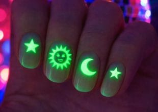 Four fingernails glowing under UV light, each with a neon green design: a star, a smiling sun, a crescent moon, and another star, against a colourful, blurred background.
