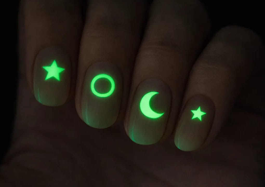 A close-up of four fingernails painted with glow-in-the-dark designs: a star, a circle, a crescent moon, and another star, all glowing green in the dark.