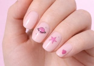 En hånd med blekrosa neglelakk har romfartstema på tre negler: en rosa planet med en ring, en rosa stjerne og et rosa hjerte. Bakgrunnen er en myk, lys rosa farge.