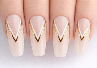 Close-up of four fingers with long, almond-shaped nails, painted in a nude colour and decorated with elegant white and gold V-shaped geometric lines near the tips.