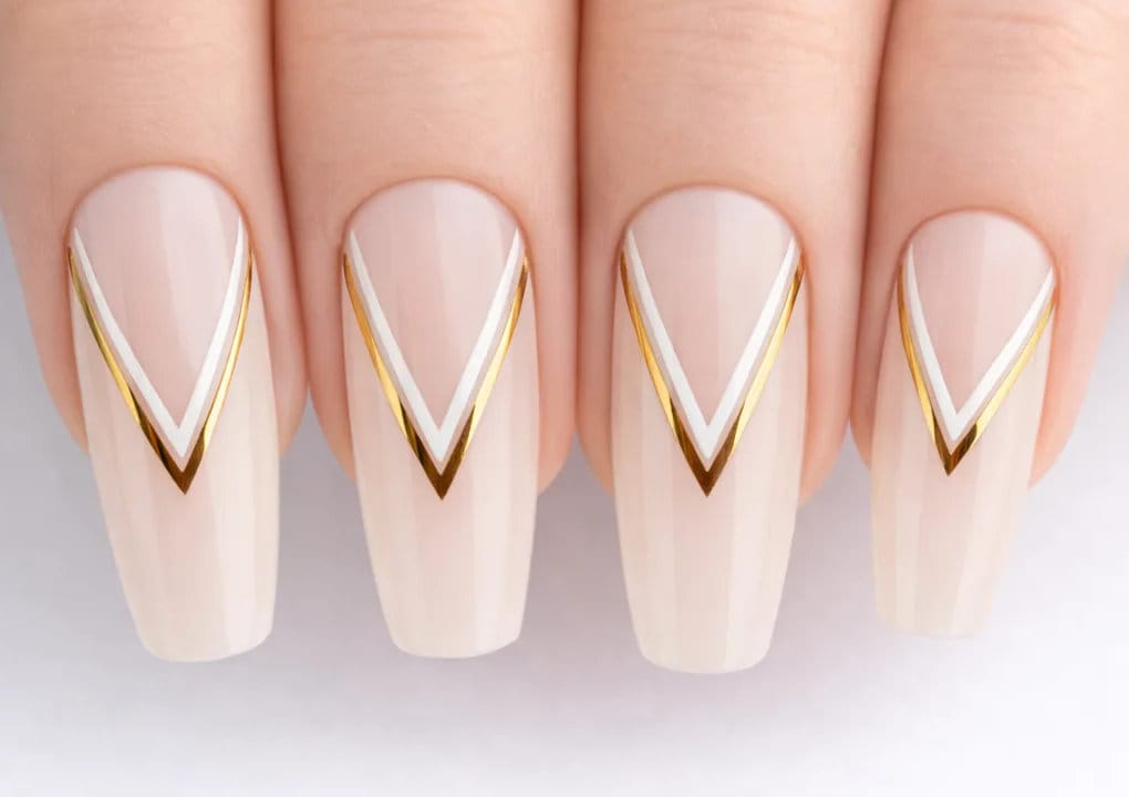 A close-up of four fingernails with a nude base color, each featuring a chevron design in white and gold lines near the tips. The nails are long and neatly manicured.