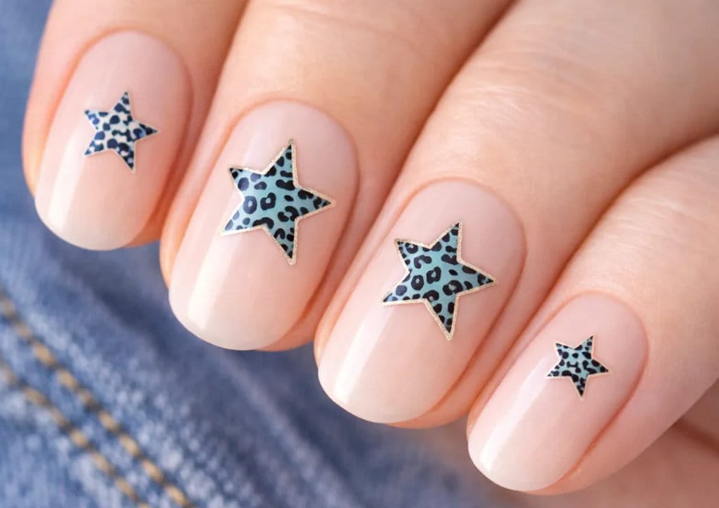 A close-up of manicured fingernails painted with a pale nude polish, each featuring a blue and black leopard print star-shaped design, with part of denim fabric visible in the background.