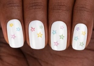 A close-up of a hand with white nail varnish decorated with small, colourful star designs in pink, yellow, green, blue, and purple on short, neatly manicured nails.