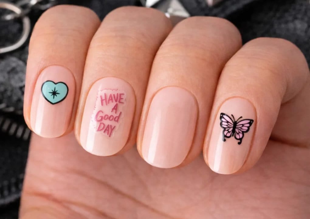 A hand with light pink nail polish; the index finger has a blue heart with a star, the middle finger says “Have a good day” in pink, and the ring finger has a purple butterfly design.