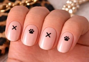 A hand with short, rounded nails painted in light pink varnish features simple black nail art: crosses on the index and ring fingers, and paw prints on the middle and little fingers.