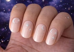 A hand with nude-coloured nails features delicate silver crescent moons and star designs, with a starry night sky background.