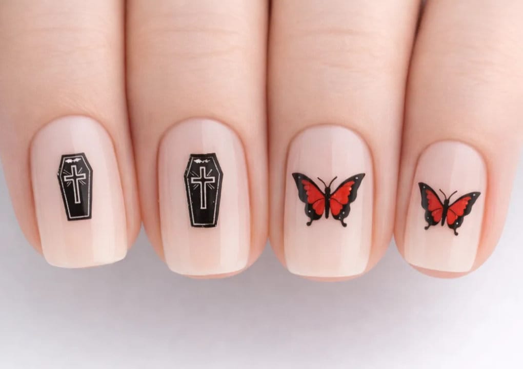 Close-up of fingernails with a nude base; two nails feature black coffin designs with white crosses, and two nails have red and black butterfly designs.