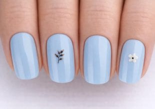 A hand with light blue polished nails; the middle nail has a small brown twig design, and the ring nail has a tiny white flower. The nails are neatly manicured with a glossy finish.