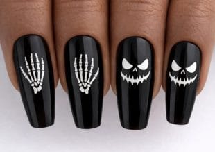 Four fingernails painted black with Hallowe’en designs: two nails have white skeleton hands and two have white scary jack-o’-lantern faces. The designs are bold and stand out against the glossy black varnish.