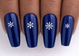 Close-up of four fingernails painted dark blue, each featuring a white snowflake design in the centre. The nails are long and have a glossy finish.