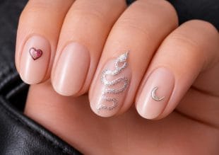 A hand with nude nail varnish features nail art: a small red heart on the little finger, a silver glitter snake on the middle finger, and a silver crescent moon on the ring finger.